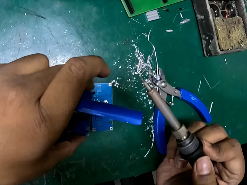 Head-camera view of a desoldering pump pressed to a circuit board while the other hand holds a soldering iron beside a pair of pliers.