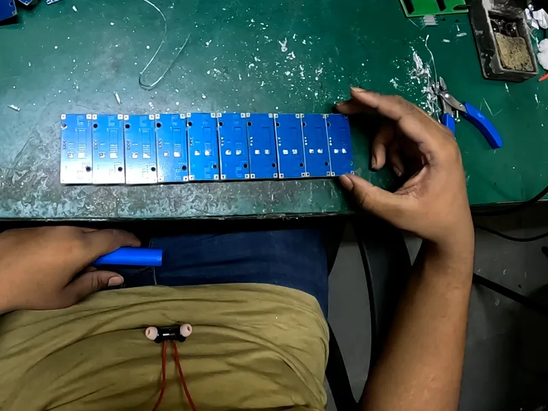 Head-camera view of a worker's right hand squaring up a row of nine blue circuit modules on a green workbench.